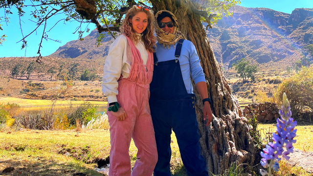 Two people in overalls standing in front of a tree with mountains in the background